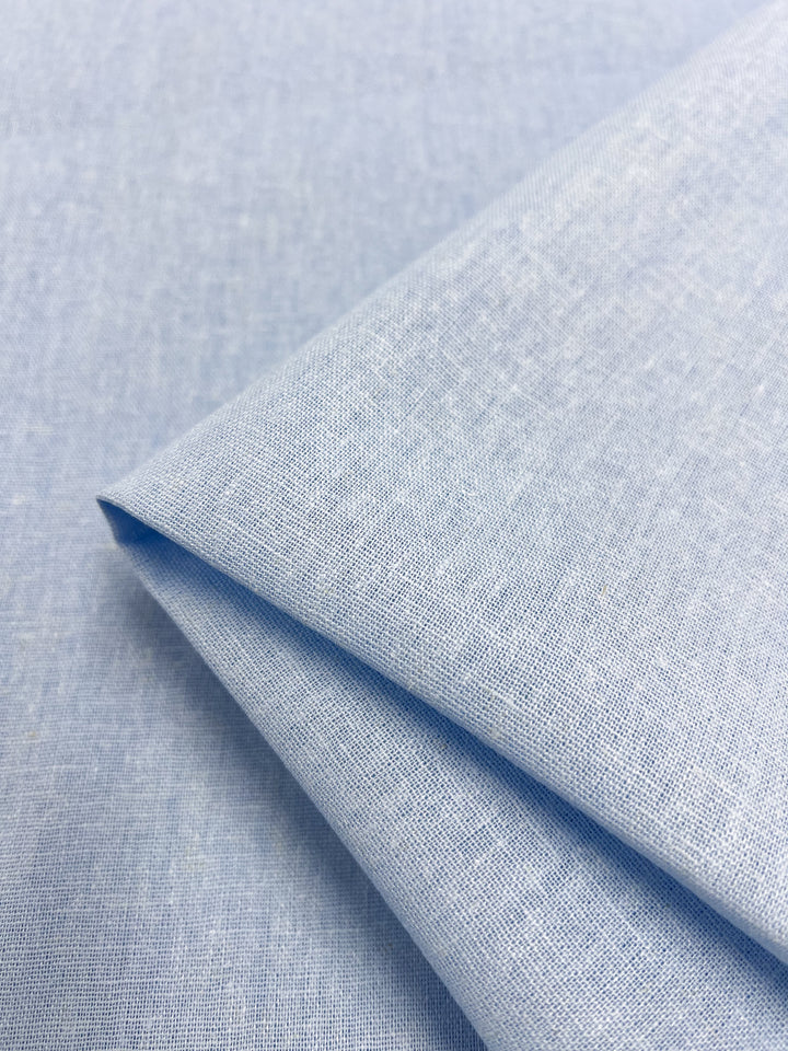 A close-up of the Premium Linen Cotton - Clear Sky - 140cm fabric from Super Cheap Fabrics shows its lightweight, light blue linen with a subtle texture. The material is folded neatly, highlighting a soft and smooth appearance reminiscent of a clear sky, perfect for clothing or home decor.