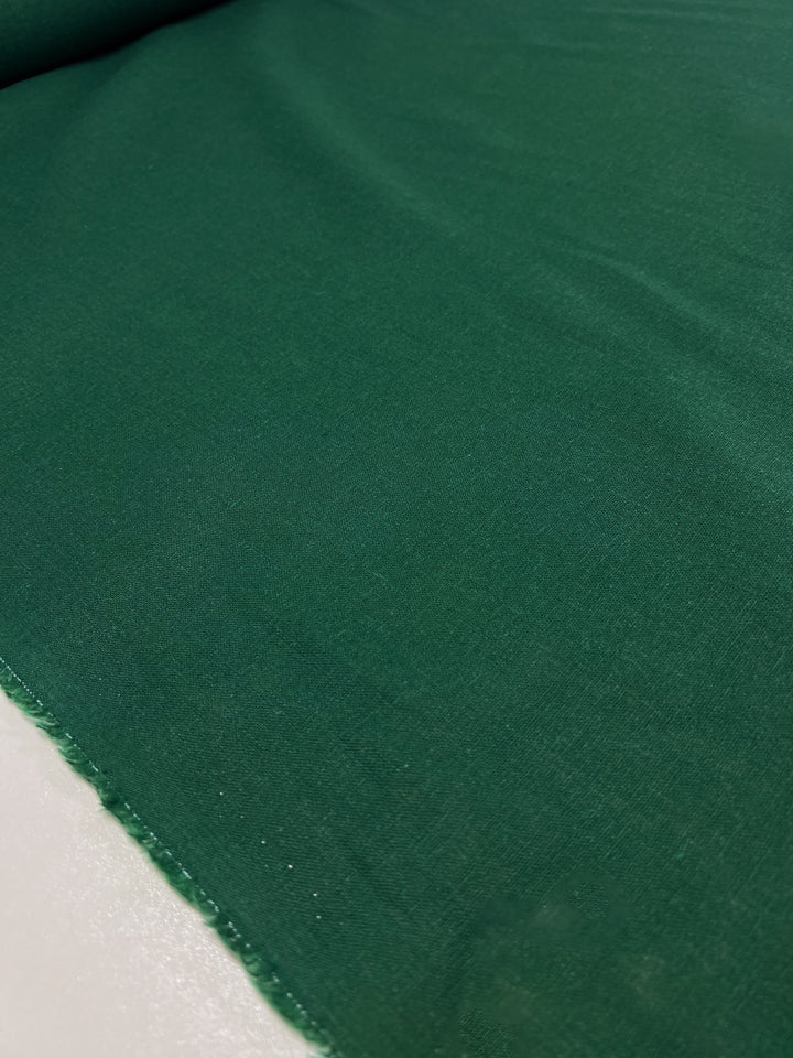 The "Premium Linen Cotton - Aventurine - 140cm" by Super Cheap Fabrics is laid out on a surface, featuring a deep aventurine hue with a smooth texture highlighted by gentle creases and lightly frayed edges.