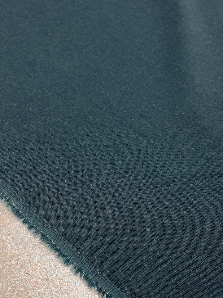 A close-up of the Premium Linen Cotton - Balsam from Super Cheap Fabrics reveals its dark green, balsam hue with a subtly textured surface, stretching diagonally from the bottom left to the top right. The fabric's fringed border is visible against a softly blurred neutral backdrop.