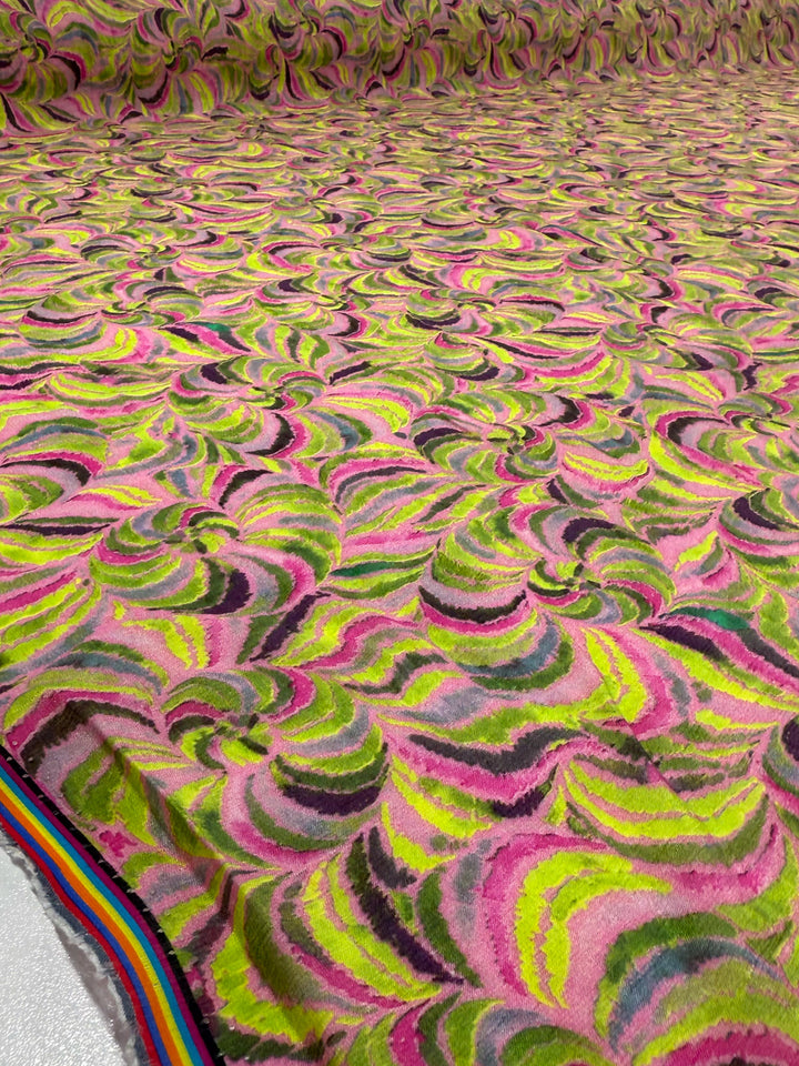 The Super Cheap Fabrics Textured Designer Cotton - Neon Vortex (145cm) features lightweight cotton with swirling marbled patterns in pink, green, yellow, brown, and black, plus a rainbow-striped edge in one corner.