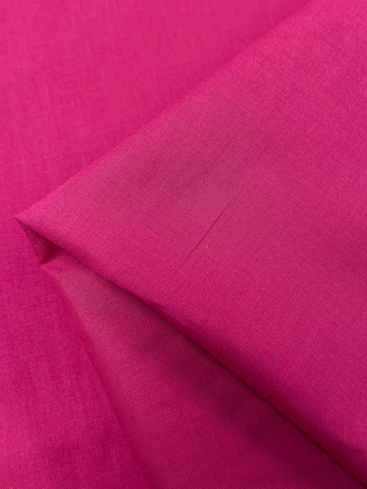 A close-up of the Super Cheap Fabrics' Cotton Voile in Fuchsia (145cm), folded neatly at one corner, showcases its smooth texture. The vibrant cotton captures light and shadow beautifully, highlighting its exquisite material quality.