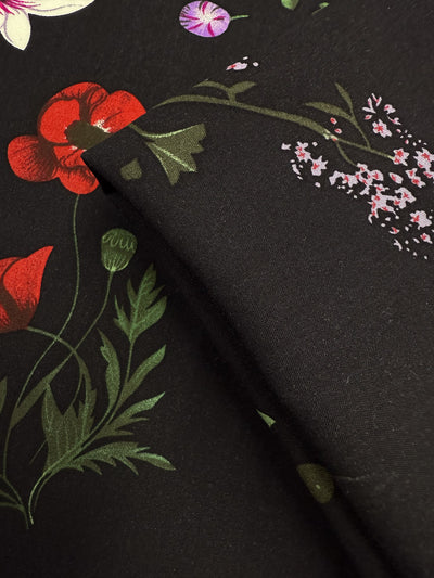 A close-up of Super Cheap Fabrics' Designer Rayon - Dark Orchard Bloom (145cm), showcasing its lightweight feel and vibrant floral print with red poppies, green leaves, and pink flowers. Fabric is partially folded to display the pattern from various angles.