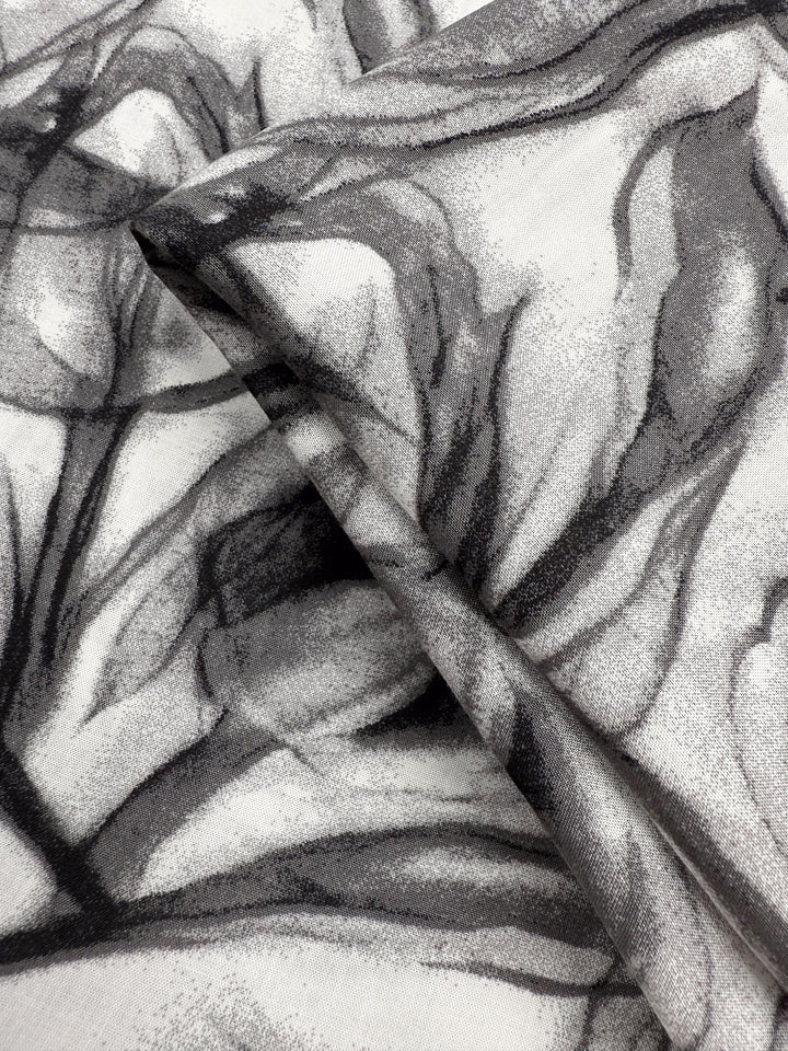 A close-up of Super Cheap Fabrics’ Designer Rayon - Shadow Petal (148cm), showing two overlapping pieces with a black and gray abstract floral print on white, featuring soft, flowing lines for a textured look.