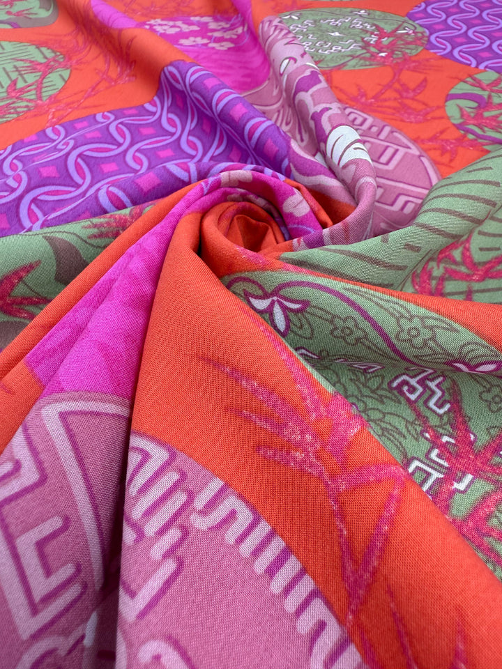 A close-up of Designer Rayon - Kyoto Lanterns (146cm) by Super Cheap Fabrics, showcasing vibrant pink, orange, and green hues enhanced with bold geometric and floral patterns in swirling folds.