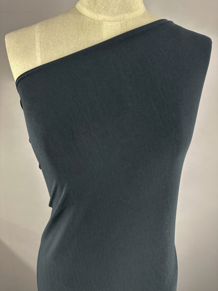 A dress form draped in a single-shoulder ITY Knit - Charcoal - 150cm fabric by Super Cheap Fabrics. The medium weight fabric covers the entire torso, extending from above the right shoulder down to below the hips, leaving the left shoulder and part of the upper chest exposed. The background is plain and light-colored.