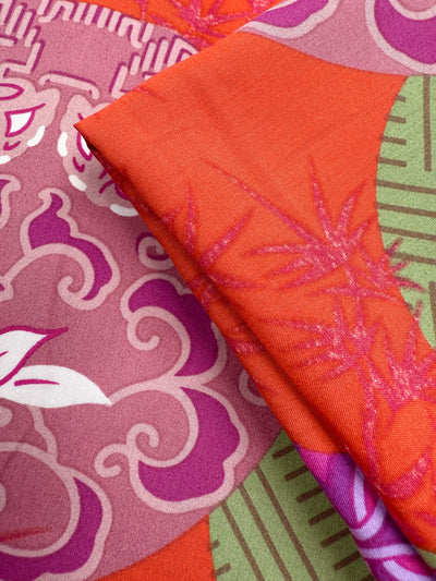 Close-up of Designer Rayon - Kyoto Lanterns (146cm) by Super Cheap Fabrics, showing folded fabric with vibrant red, orange, and pink patterns featuring abstract cloud motifs, bamboo leaves, and green and white geometric shapes.