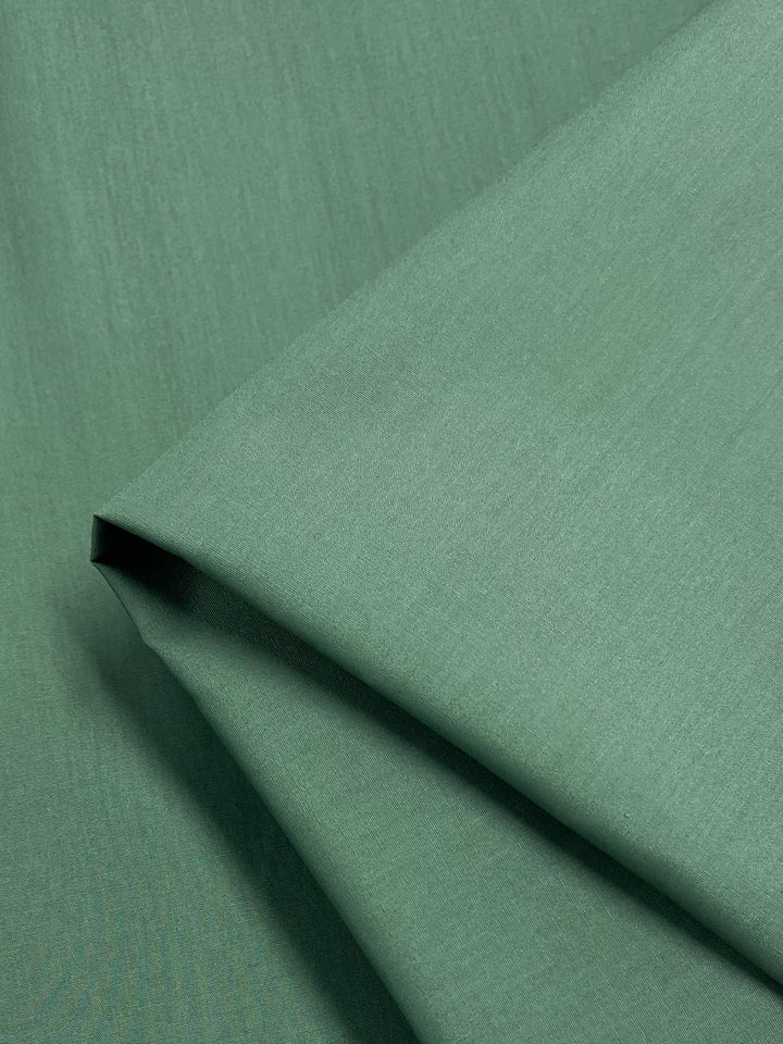 A close-up of lightweight, folded Loden Frost poplin from Super Cheap Fabrics highlights its smooth texture and even coloring. Neatly arranged with clean lines, the fabric captures a subtle play of light and shadow—ideal for crafting durable childrenswear. Measures 148cm wide.