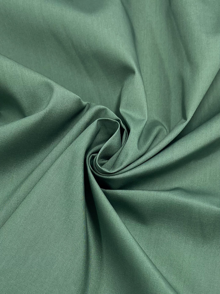 A detailed shot of Poplin - Loden Frost by Super Cheap Fabrics, showcasing its soft, lightweight poly/cotton blend with a smooth texture. Intricately folded to form a spiral, its gentle sheen enriches the vibrant green hue—ideal for childrenswear. Width: 148cm.