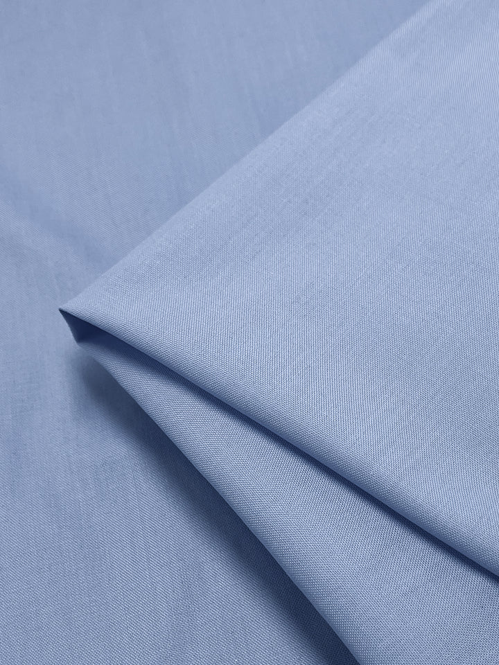 A close-up of Super Cheap Fabrics' "Oxford Poplin - Vista Blue - 148cm," a light blue polyester-cotton blend, displays its smooth, slightly textured surface. The non-stretch material is folded neatly, highlighting its soft texture and subtle sheen.