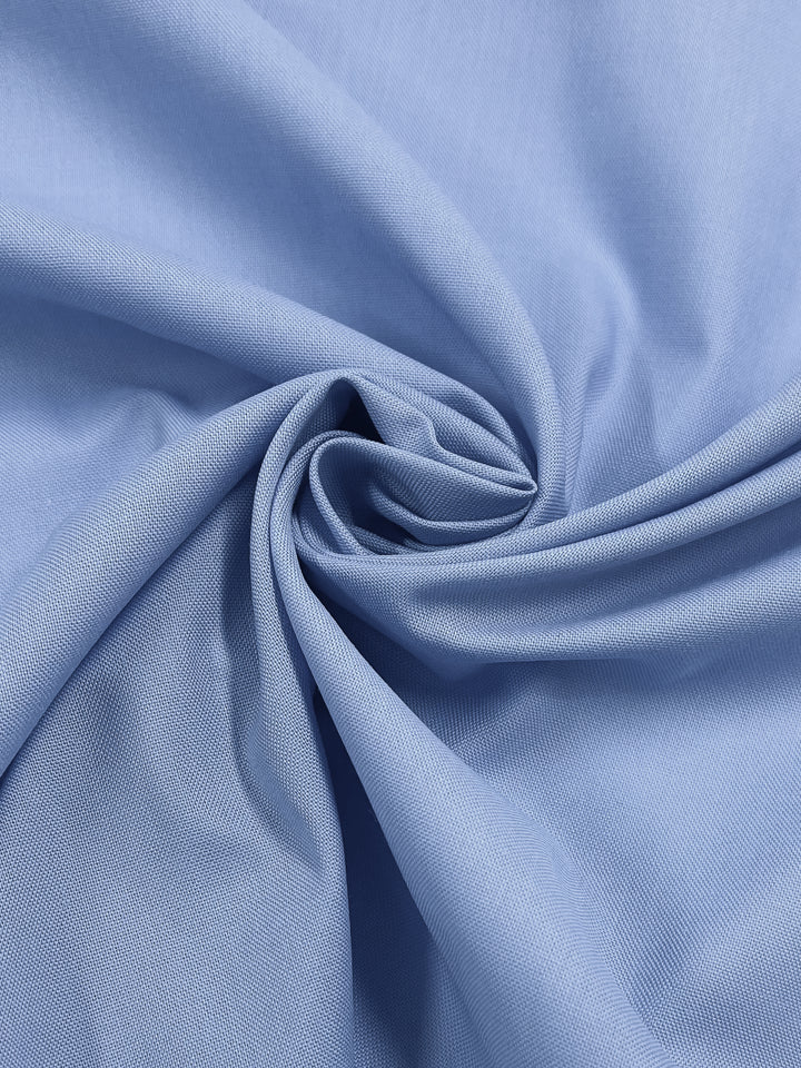 The Oxford Poplin - Vista Blue by Super Cheap Fabrics features a close-up of the light blue, non-stretch fabric spiraled. Its smooth, slightly textured surface forms soft, flowing patterns with elegant folds and curves on its 148cm wide material.