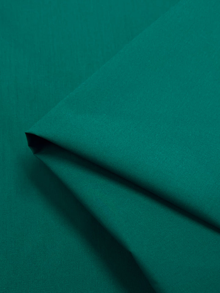 A close-up of Poplin - Kayaking fabric by Super Cheap Fabrics displays its dark green polyester cotton blend, neatly folded to reveal a textured, smooth surface forming a triangular shape on the right.