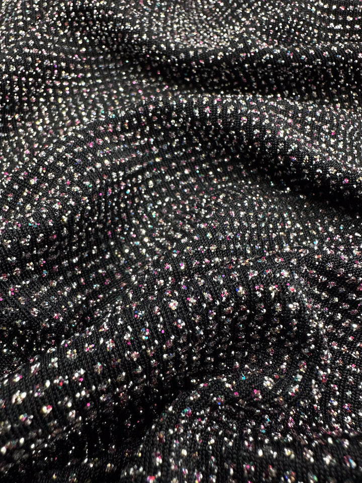 A close-up of Metallic Lycra - Disco - 150cm from Super Cheap Fabrics shows its textured black surface with reflective multi-color specks, creating a sparkling effect. The ribbed pattern enhances its soft, luxurious feel, ideal for crafting elegant dresses.