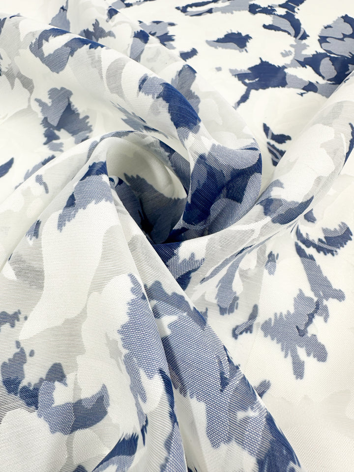 A close-up of the Deluxe Print - Blue Flora - 150cm by Super Cheap Fabrics, showcasing its lightweight polyester fabric with an abstract floral print in blue and gray, elegantly draped to highlight texture and pattern.