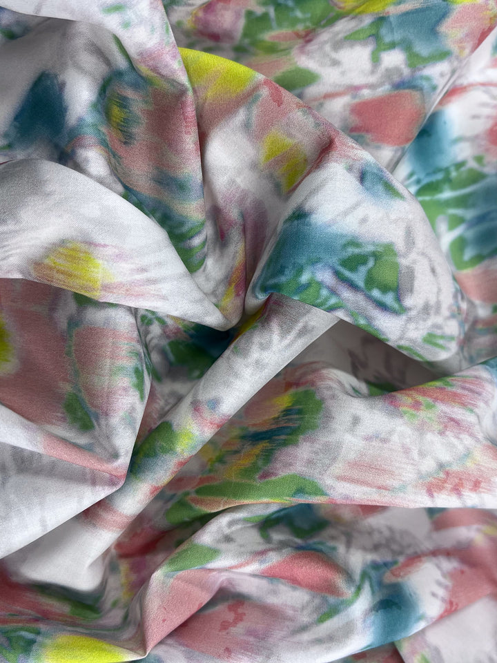 A close-up view of Super Cheap Fabrics' Printed Cotton - Terrain - 150cm showcases its lightweight cotton fabric with a colorful abstract pattern in pastel blues, pinks, yellows, and greens. The natural fibers are gently folded for a textured and flowing look.