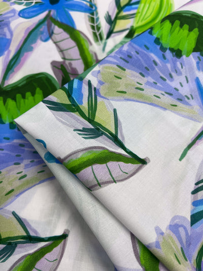 Close-up of Super Cheap Fabrics' Printed Cotton - Blue Paradise. This lightweight fabric features a vibrant floral pattern in blue, green, and purple with abstract leaves and petals. Its natural cotton fibers enhance the soft folds of its lively texture. Width: 150cm.