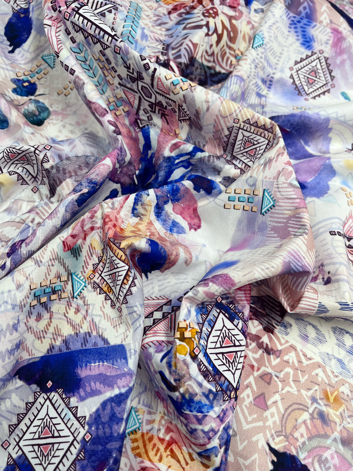 A close-up of the Super Cheap Fabrics Printed Cotton - Digital - 150cm, featuring an intricate design with geometric shapes and abstract leaf motifs in blue, purple, pink, and gold on a light background. This lightweight cotton is expertly gathered and folded.