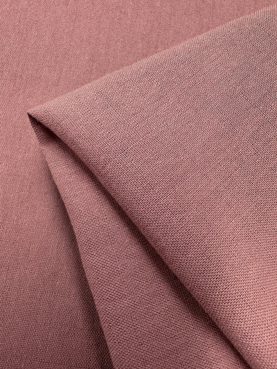 A close-up of a folded piece of Super Cheap Fabrics Linen Blend - Old Rose - 147cm with a slightly textured surface. The fabric appears soft and smooth, with visible weave patterns. The folds create gentle shadows, giving a sense of depth and material quality, showcasing its old rose hue.