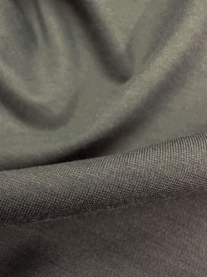 Close-up of dark gray Ponte De Roma fabric, "Deep Depths," by Super Cheap Fabrics. This Australia-sourced material has a smooth, slightly shiny texture with visible weave, creating gentle curves and shadows, emphasizing its soft, flexible 4-way stretch. Width: 155cm.