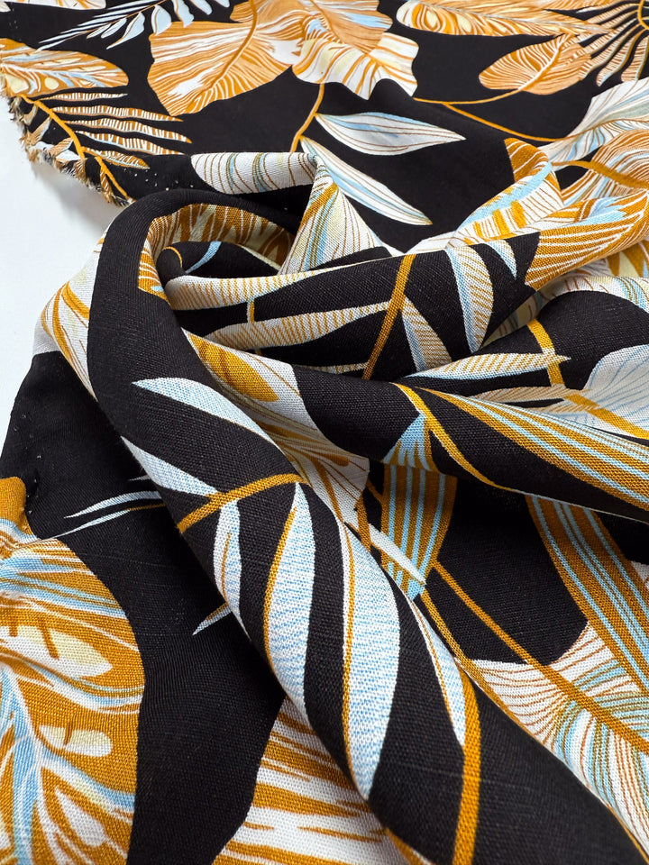 A close-up of Super Cheap Fabrics’ Bamboo Rayon - Tropic Gold (150cm), featuring lightweight bamboo rayon with a bold tropical leaf print in mustard yellow, white, and light blue on black, draped in soft folds.