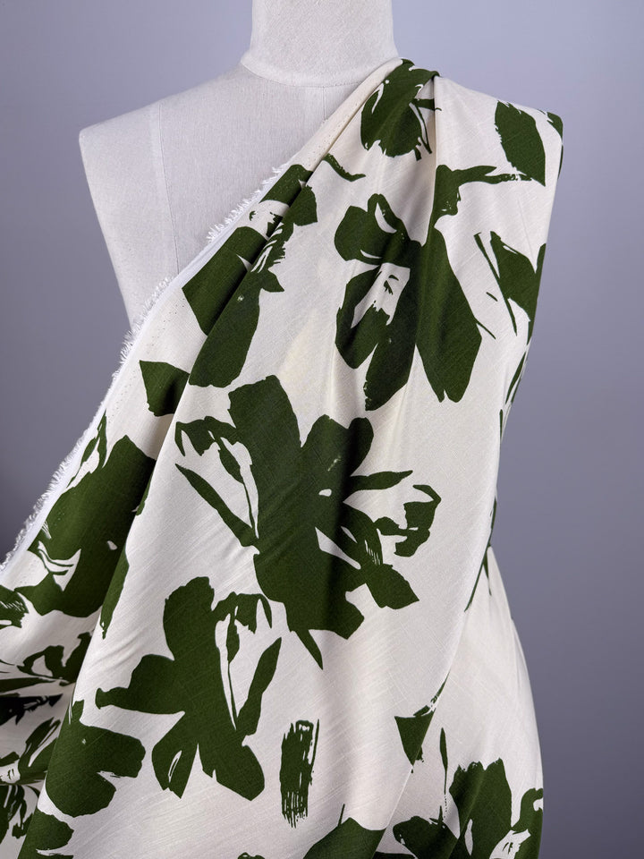 The Bamboo Rayon - Olive Bloom (150cm) from Super Cheap Fabrics features lightweight, off-white fabric with bold green leaf patterns—ideal for beginner projects—shown on a dress form against a light gray background.