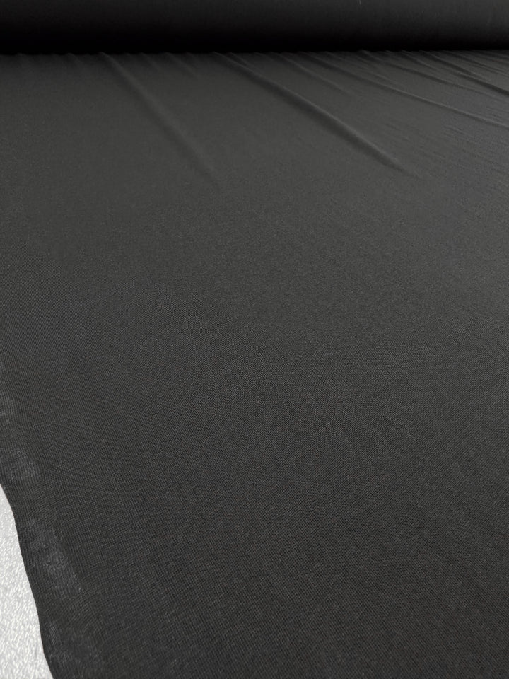 A large roll of Super Cheap Fabrics' Lightweight Cotton Jersey - Black - 145cm unfurls smoothly on a flat surface. The subtle textures are visible under lighting, showcasing its lightweight yet consistent quality in a seamless, uniform sheet.