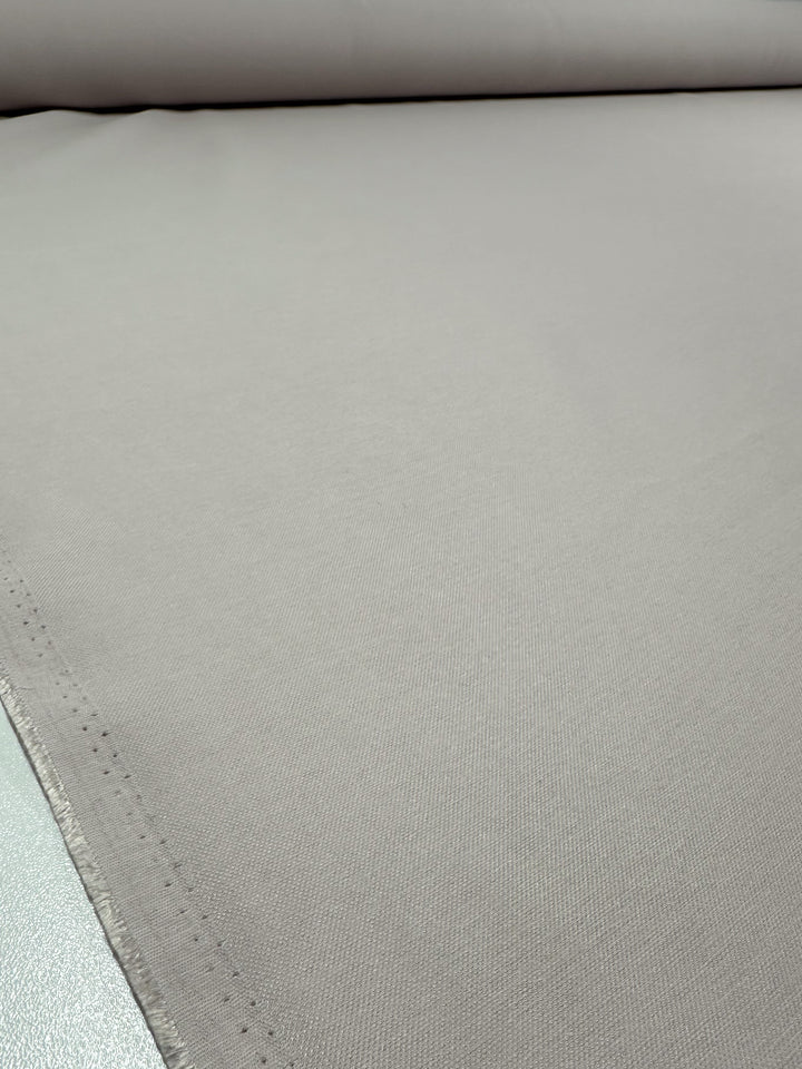 A close-up of Super Cheap Fabrics' "Suiting - Lilac Ash - 144cm," a smooth, light grey suiting fabric with a subtle texture, showcases its light to medium weight material, even surface, and slightly frayed edge at the bottom left corner.