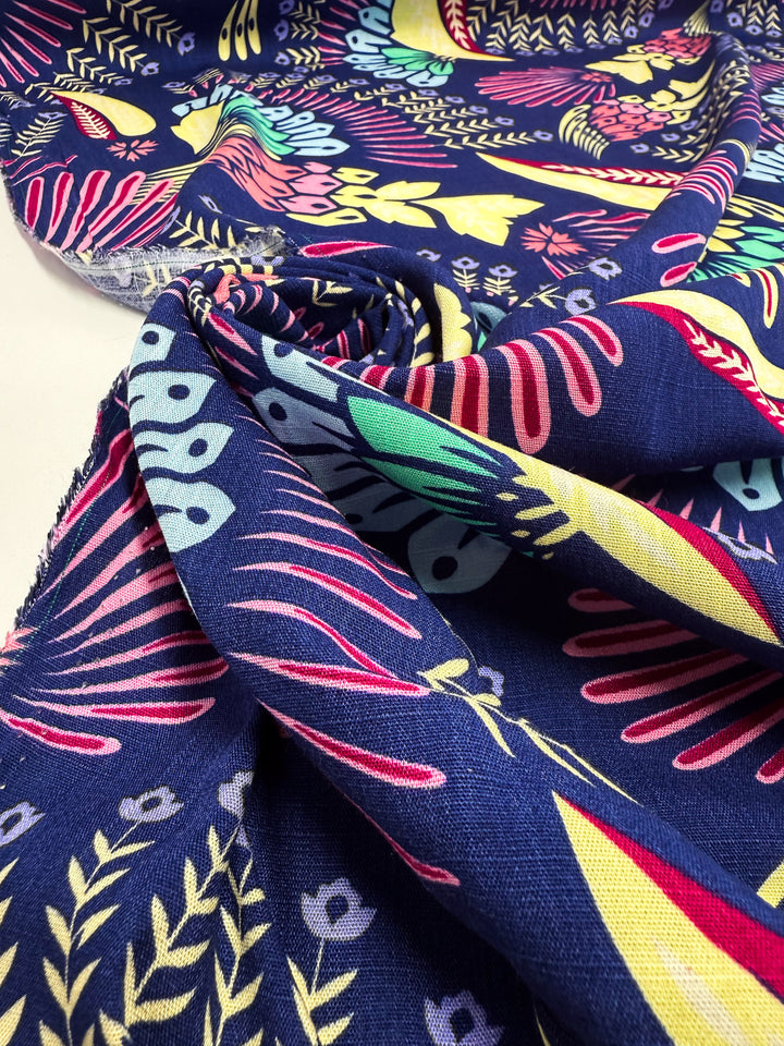 A close-up of Super Cheap Fabrics' Bamboo Rayon - Midnight Carnival (150cm) shows lightweight, softly folded dark blue fabric with a vivid pink, yellow, red, and light blue floral and leaf pattern.