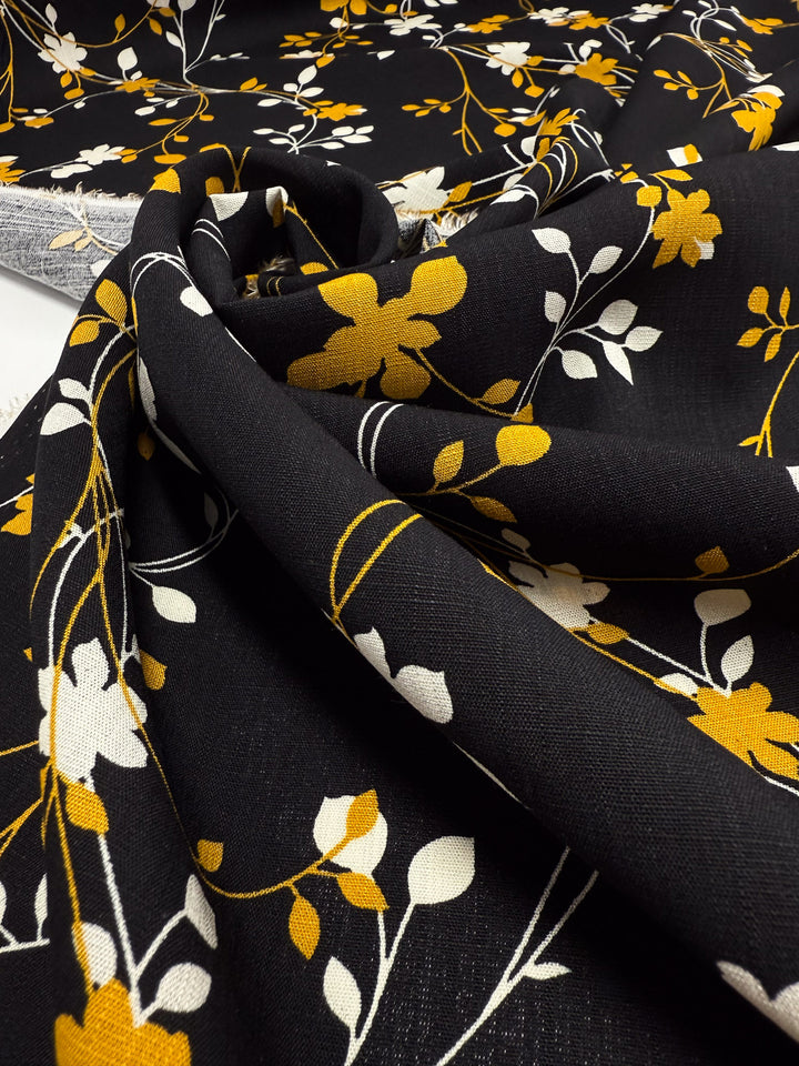 Super Cheap Fabrics' Bamboo Rayon - Golden Vine (150cm) features a lightweight black fabric with yellow and white floral patterns and white stems, shown draped to highlight its texture—ideal for beginner-friendly sewing projects.