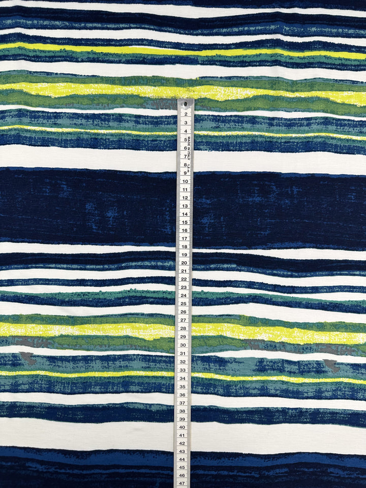 The Printed Lycra - Ocean Stripe from Super Cheap Fabrics, with its blue, green, yellow, and white horizontal stripes on Polyester/Spandex fabric, evokes summer vibes. A vertically placed measuring tape shows measurements in centimeters across the 145cm width.