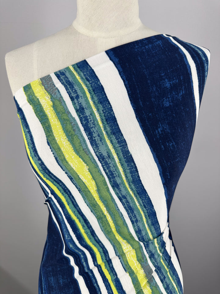 Displayed on a mannequin, the dress showcases a one-shoulder Printed Lycra design in Ocean Stripe colors of blue, white, yellow, and green. Made by Super Cheap Fabrics with a Polyester/Spandex blend, it offers a textured look that enhances its vibrant summer pattern.