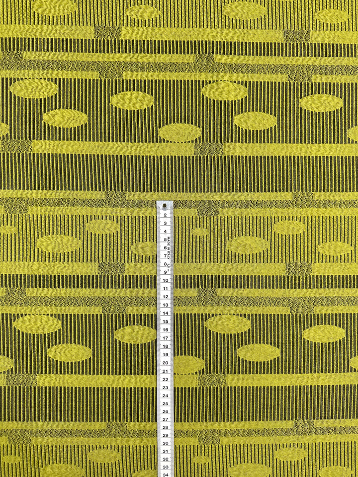 The Jacquard Knit - Chartreuse by Super Cheap Fabrics, measuring 138cm wide, showcases a yellow and dark green geometric pattern with vertical stripes and circles. A centrally placed measuring tape marked from 1 to 31 emphasizes its textured design for style and stretchable comfort.