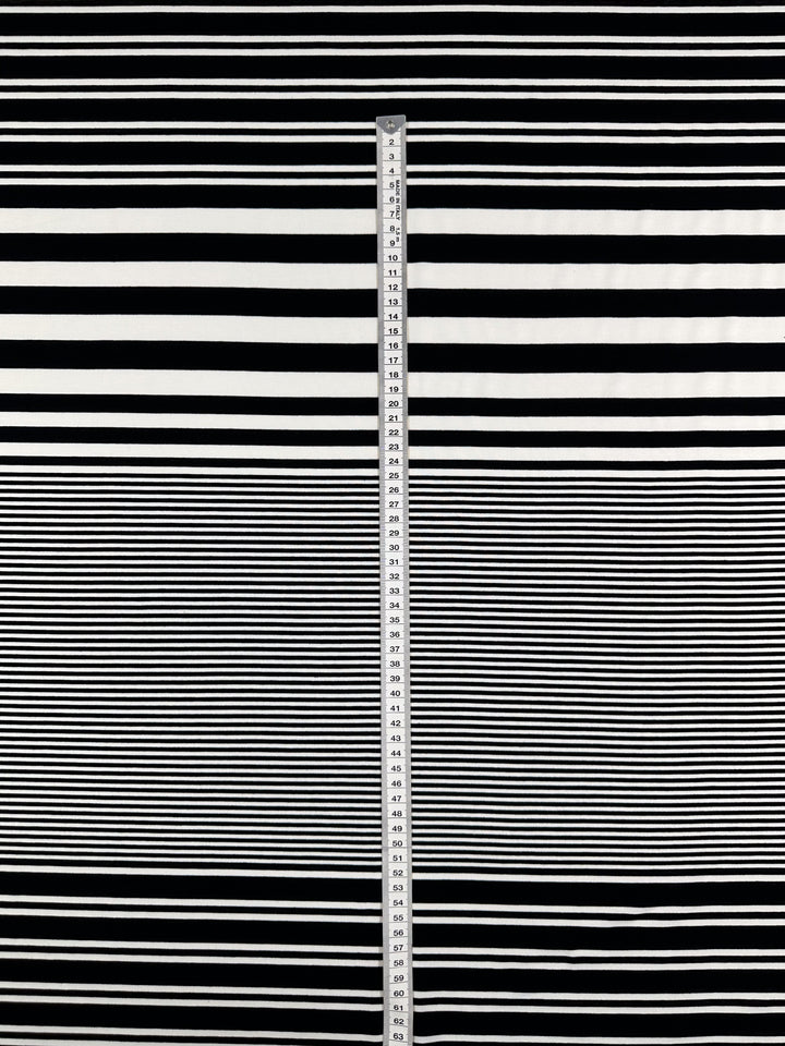 A measuring stick stands against alternating black and white stripes, resembling the soft, stretchy Viscose Jersey - Paralello fabric from Super Cheap Fabrics, which drapes gracefully with thicker stripes at the top and thinner ones below for a fluid effect.