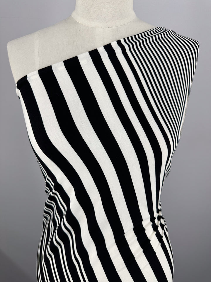 A mannequin displays a black and white striped, one-shoulder garment made of Viscose Jersey - Paralello from Super Cheap Fabrics. The stretchy fabric's asymmetrical stripes form a striking pattern against a plain gray background.