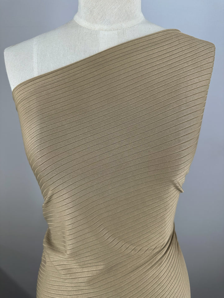 A close-up of the Rib Knit - Dune - 168cm by Super Cheap Fabrics showcases its beige, ribbed, one-shoulder design on a mannequin. The medium-weight, stretchy fabric drapes elegantly over the shoulder with a textured knit. A neutral gray backdrop highlights its intricate details.