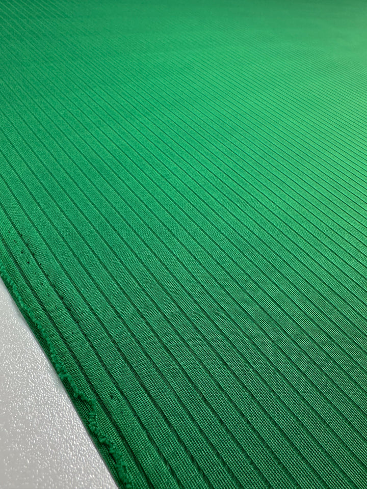 A close-up of the Rib Knit - Abundant Green fabric from Super Cheap Fabrics shows vibrant medium-weight green with diagonal thin ridges. The smooth and slightly shiny texture enhances its pattern, with the edge visible on the left side. Width: 177cm.