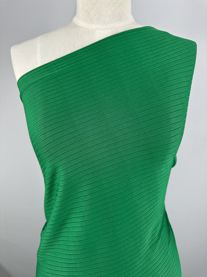 A mannequin displays a one-shoulder dress made from Super Cheap Fabrics' Rib Knit in Abundant Green (177cm), featuring a textured diagonal stripe pattern and crafted from medium weight rib knit fabric that delivers generous stretch for ultimate comfort, set against a plain gray background.