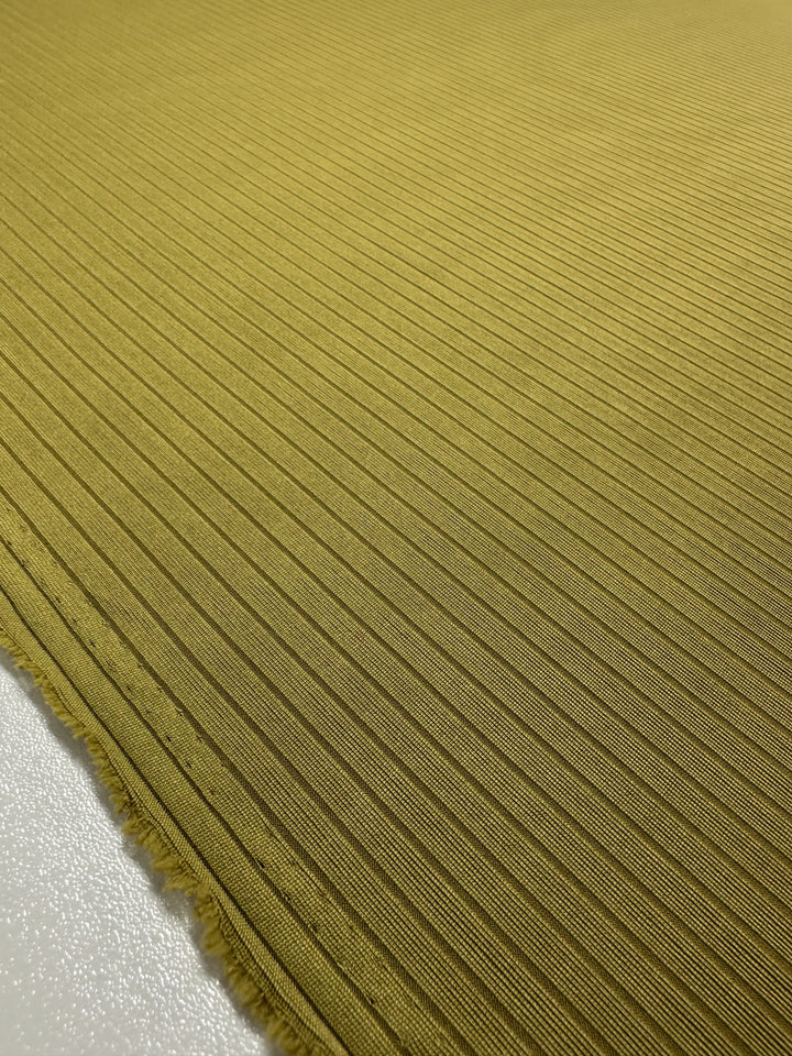 A close-up of the Super Cheap Fabrics "Rib Knit - Avocado Oil - 173cm" displays its yellow hue, thin, evenly spaced horizontal ridges, and frayed edges on the left. Laid out on a light surface, its intricate design is prominent.