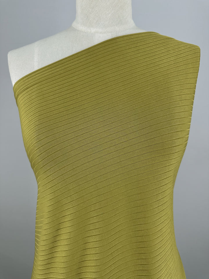 A mannequin is adorned with Super Cheap Fabrics' Rib Knit - Avocado Oil, a 173cm medium weight fabric with diagonal lines. The neutral gray backdrop accentuates its stretchy comfort and texture.