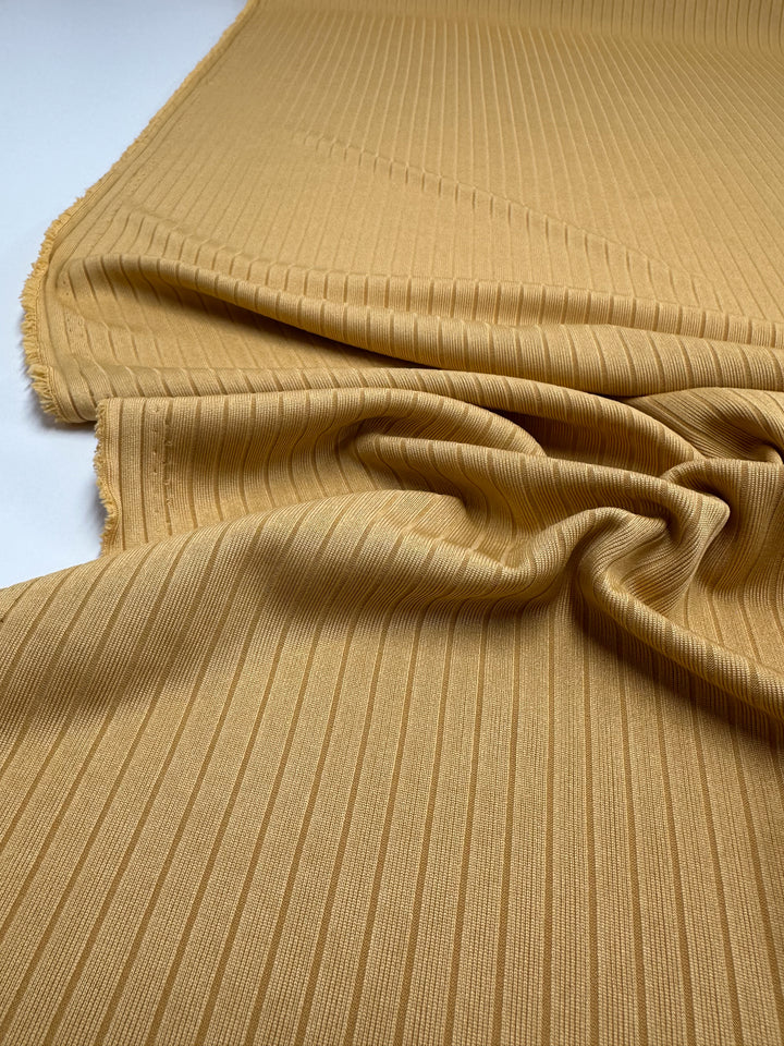 A close-up of Super Cheap Fabrics' "Rib Knit - Pale Golden Nugget - 173cm" shows its textured, medium-weight fabric with a slight center gather, highlighting its sheen. The edges appear slightly frayed against a plain, light background.