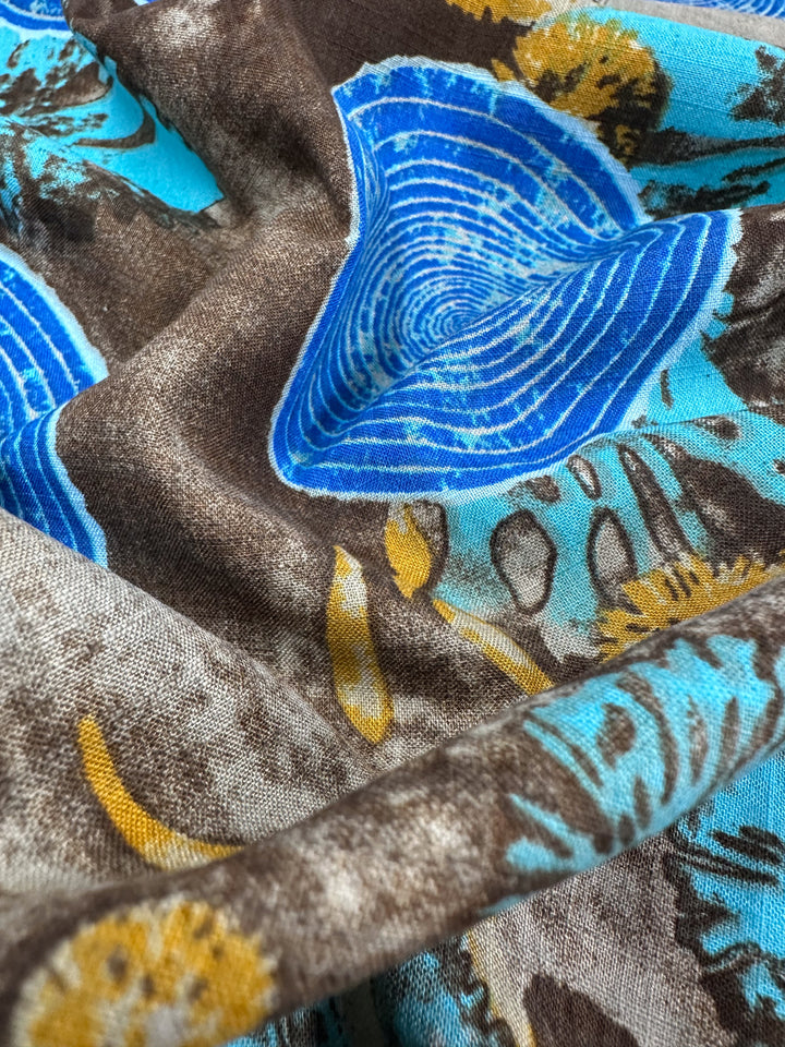 Close-up of the Linen Blend - Radar On Earth. This Super Cheap Fabrics material showcases a colorful abstract pattern with blue tree ring-like circles on a brown background, accented by hints of yellow and turquoise. The soft, textured fabric is a lightweight linen cotton blend.