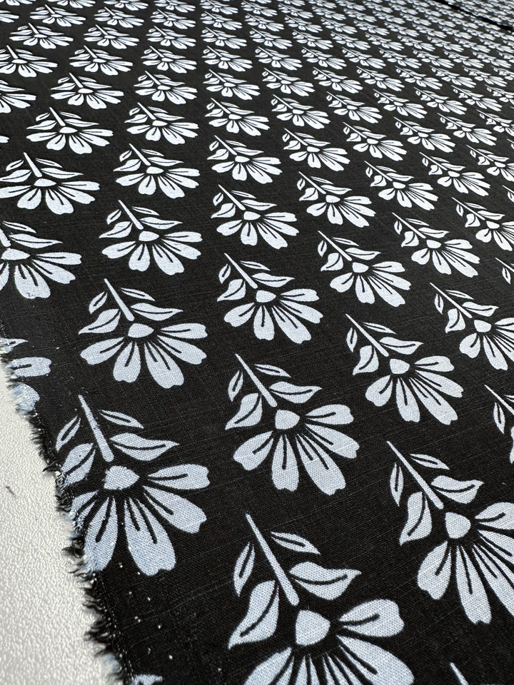 The "Chambray Floretta" by Super Cheap Fabrics is a 138cm linen blend featuring stylized gray flowers with leaves in diagonal rows on black. Its breathable fabric and softly frayed edges enhance its rustic charm.