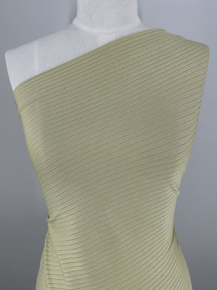 A mannequin displays a one-shoulder dress crafted from Super Cheap Fabrics' Rib Knit in Hay, 170cm, exhibiting a form-fitting silhouette and horizontal ribbing against a neutral gray backdrop.