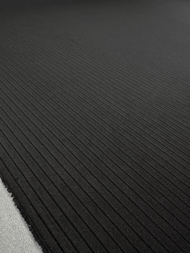 A close-up of the Super Cheap Fabrics' Rib Knit - Black - 170cm showcases a black textured fabric with narrow, diagonal ribbed lines running from top left to bottom right. The medium-weight knit looks smooth and has slightly frayed edges at the bottom.