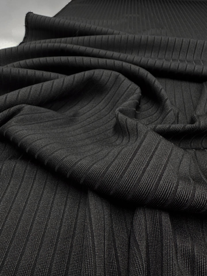 The black medium weight rib knit fabric from Super Cheap Fabrics features a visible texture and folds, forming a wavy pattern. The stretchy material is soft and slightly shiny under light, enhancing its comfort. Product: Rib Knit - Black - 170cm.