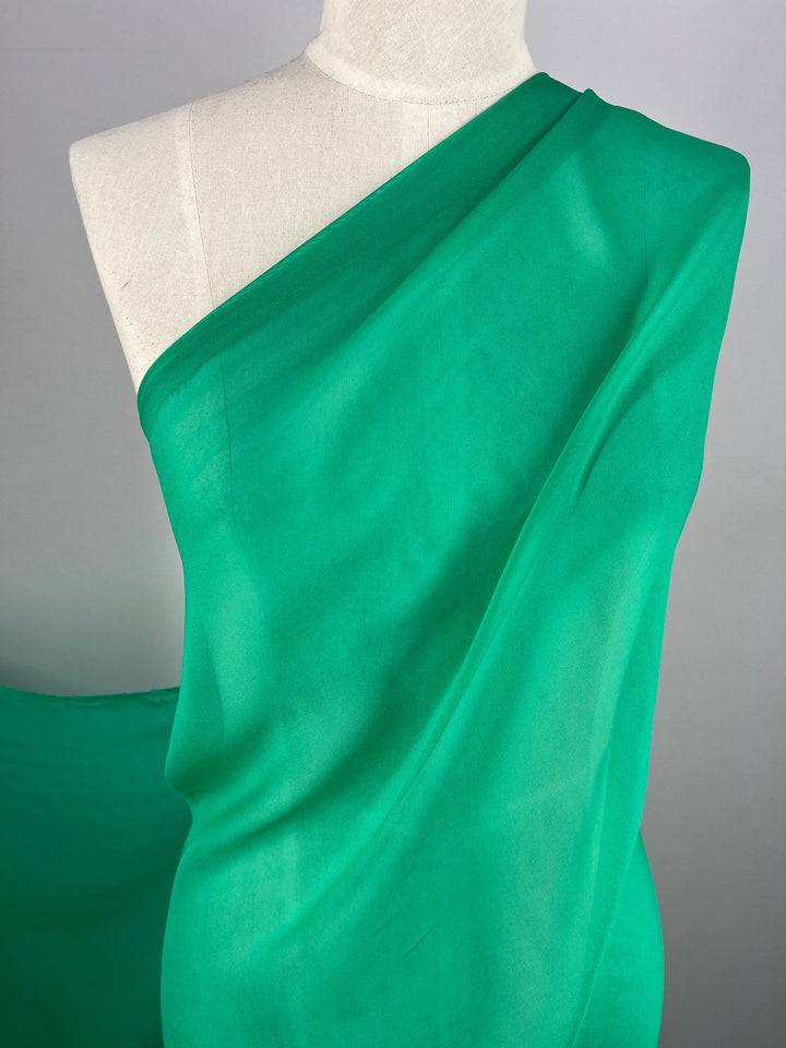 A mannequin elegantly adorned with Super Cheap Fabrics' Silk Georgette in Emerald, measuring 135cm wide, gracefully drapes the vibrant green fabric over one shoulder and wraps it around the torso. The remainder of this delicate and lightweight material trails off camera, standing out against a plain, light grey background that highlights its vivid color and exquisite texture.