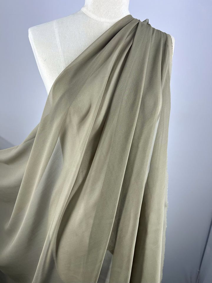 A mannequin draped in Super Cheap Fabrics' Pure Silk Georgette - Dune - 135cm, set against a white background. The olive-green fabric is lightweight, soft, and translucent, elegantly cascading over the mannequin's shoulder—ideal for spring and summer wear.