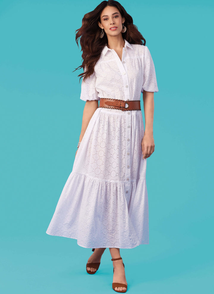 A woman stands against a blue background wearing a chic outfit inspired by the Mccall's M8285 pattern from Super Cheap Fabrics. Her ensemble features a white eyelet shirtdress with short sleeves, accessorized with a brown belt and matching strappy sandals. Her long hair flows in loose waves, complemented by hoop earrings, creating an effortlessly fashionable look.