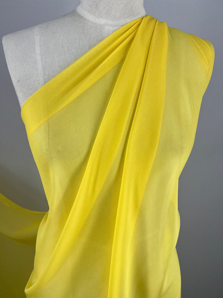 A mannequin is draped with a flowing, sheer Super Cheap Fabrics Silk Georgette in the Buttercup colorway. The fabric, measuring 135cm wide, is arranged diagonally across the chest and shoulder, creating elegant folds that accentuate its lightweight and translucent nature. Ideal for spring and summer outfits, the plain neutral background highlights the fabric’s vibrancy.