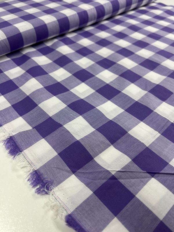 Close-up of the Cotton Gingham XL - Sand Verbena fabric by Super Cheap Fabrics, featuring a purple and white checkered pattern laid out flat with visible selvage edge. This 112cm wide natural cotton fabric showcases alternating colored squares for a classic and symmetric look, ideal for various applications.