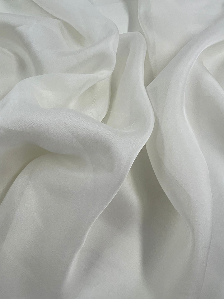 A close-up image of the Platinum Pure Silk Chiffon fabric from Super Cheap Fabrics showcases its soft, white, extra lightweight texture with gentle folds and creases. The 100% silk material appears delicate and smooth, exuding an elegant and serene quality.